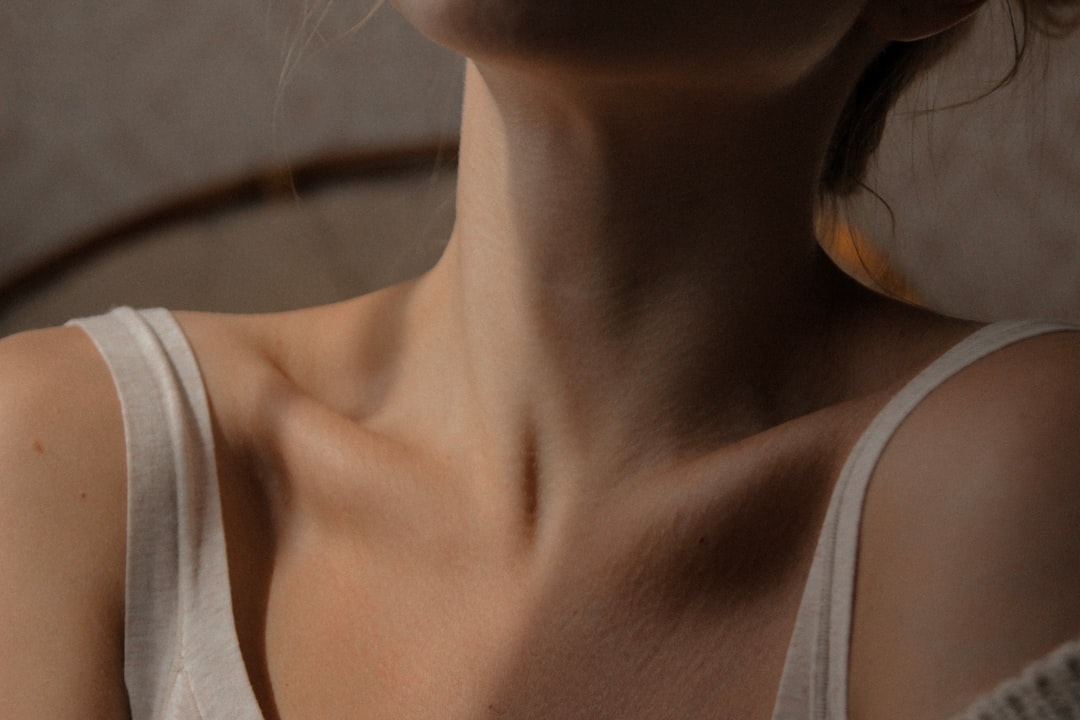 A woman in her 30s sitting near a window, soft natural light, gently touching the back of her neck with a relaxed expression, warm and reassuring atmosphere, no